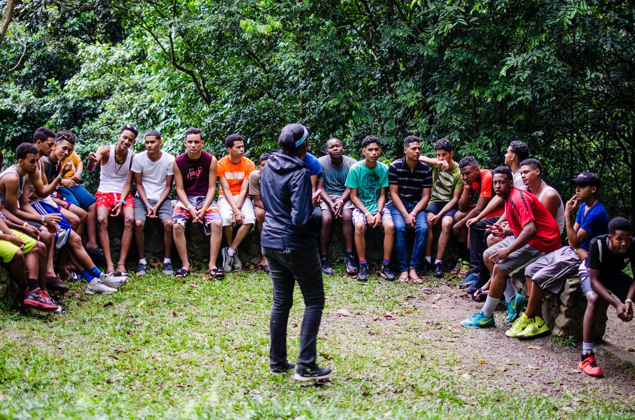 A group of teenagers and adults listen attentively during an outdoor session in a green park.