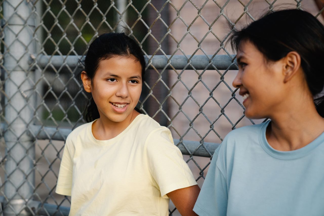 Two young women smiling and talking while leaning against a chain link fence outdoors.